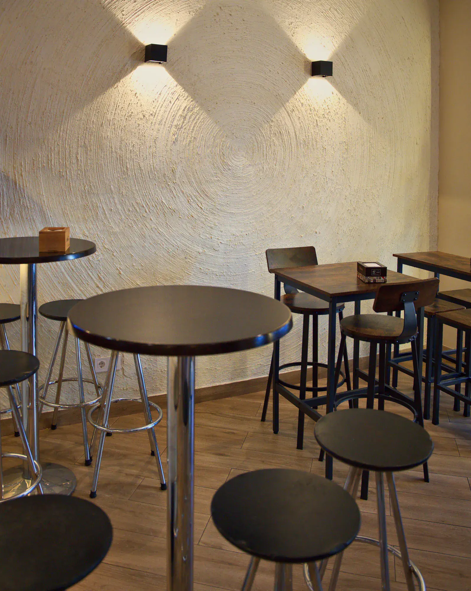 Salón de Che Pibita: mesas altas de madera, taburetes y pared blanca iluminada por apliques